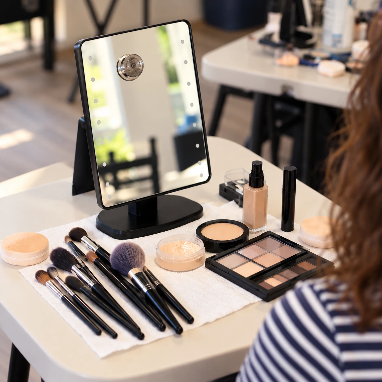 A makeup station set up with brushes, foundation, powder and an eyeshadow palette in front of a lit mirror